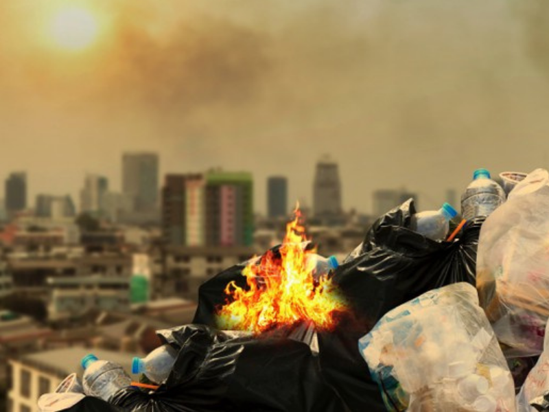 Pile of garbage bags with visible flames and smoke, with a city skyline in the background at sunset